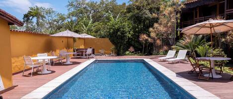A rectangular swimming pool with tiled edges, surrounded by a wooden deck. There are white lounge chairs and tables with umbrellas, and a building with a thatched roof in the background.