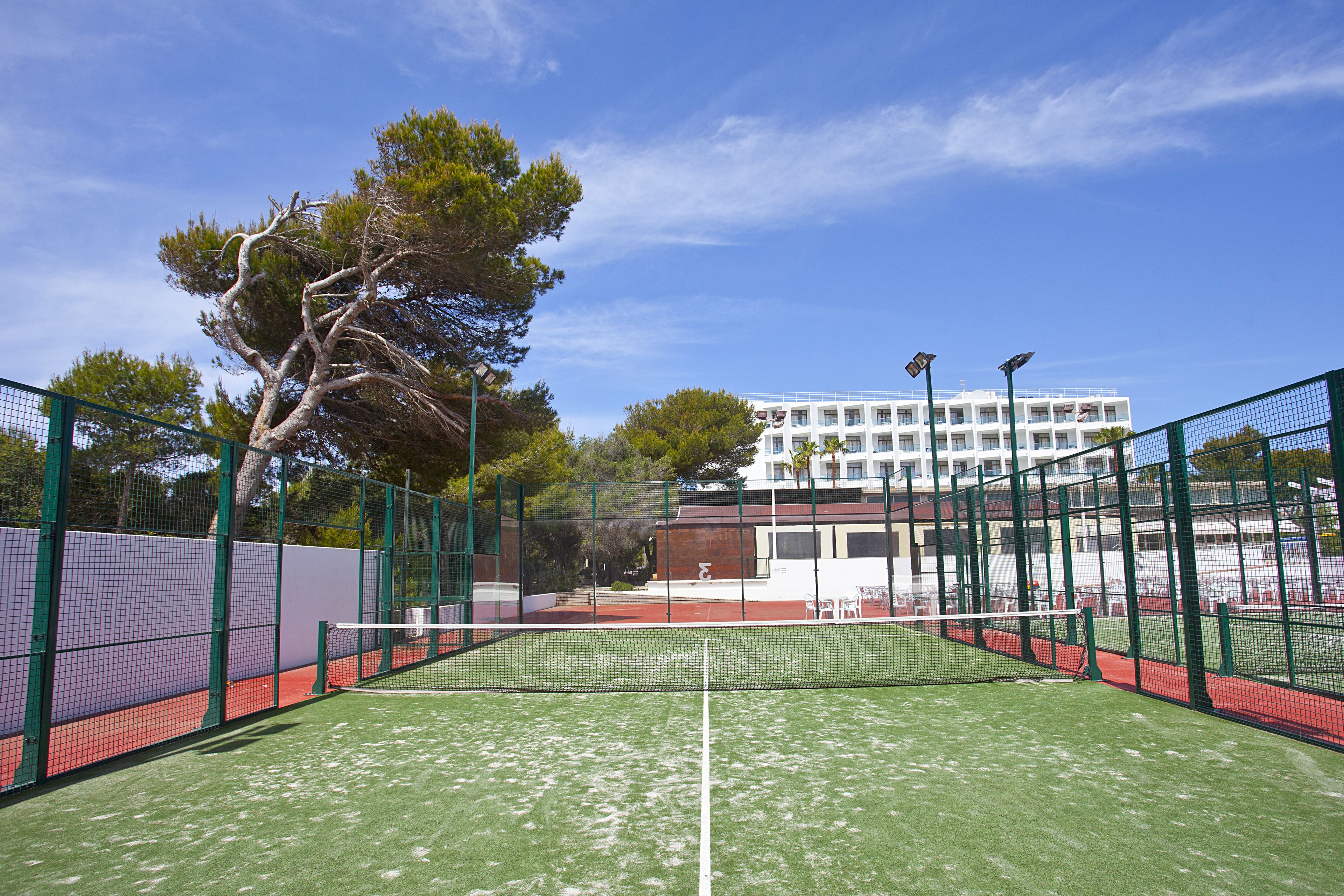 An outdoor tennis court with a net, surrounded by green fencing, and a building in the background.