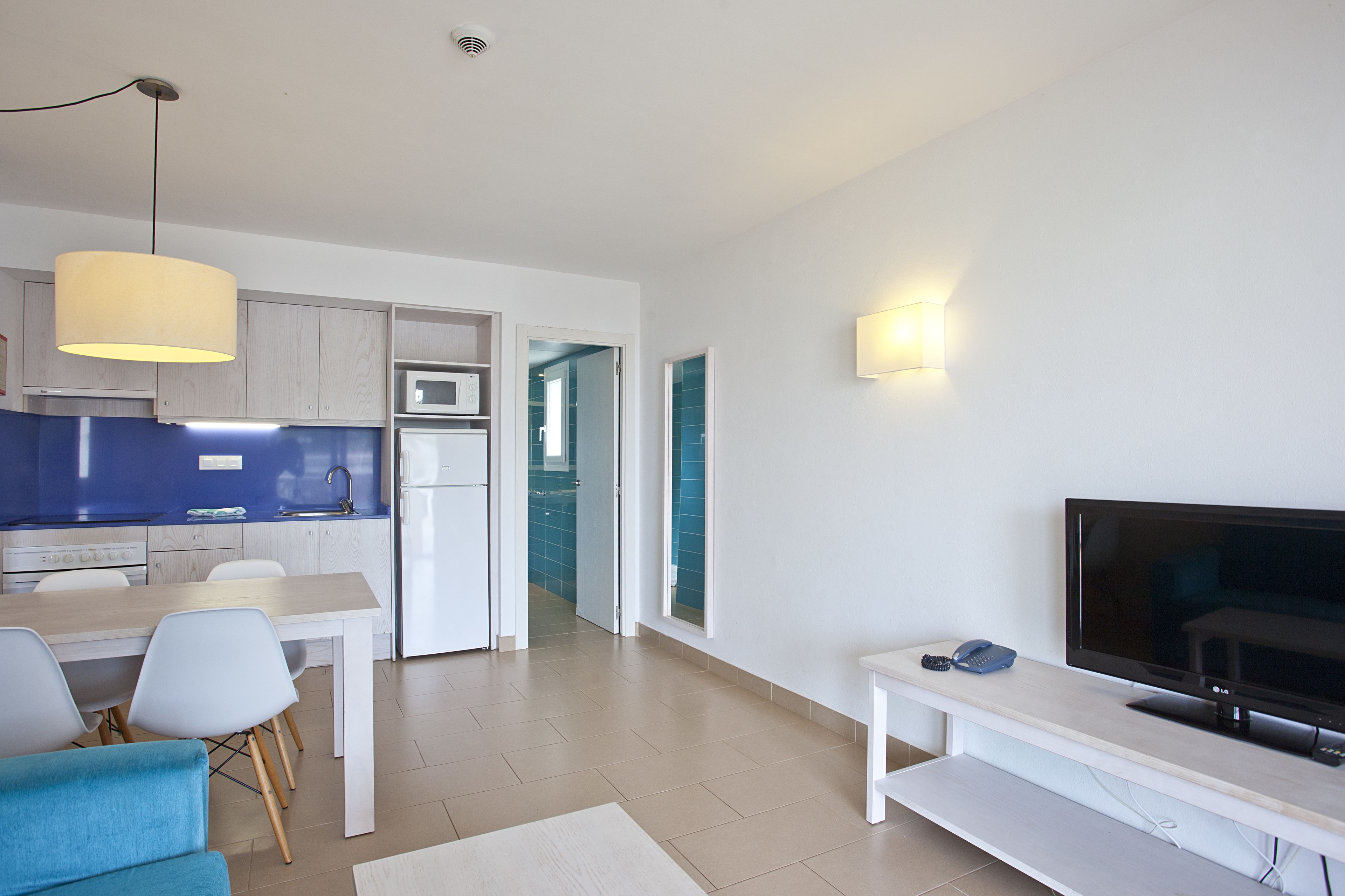 Una sala de estar moderna con televisor de pantalla plana, comedor con mesa y sillas, y cocina con armarios azules y microondas.