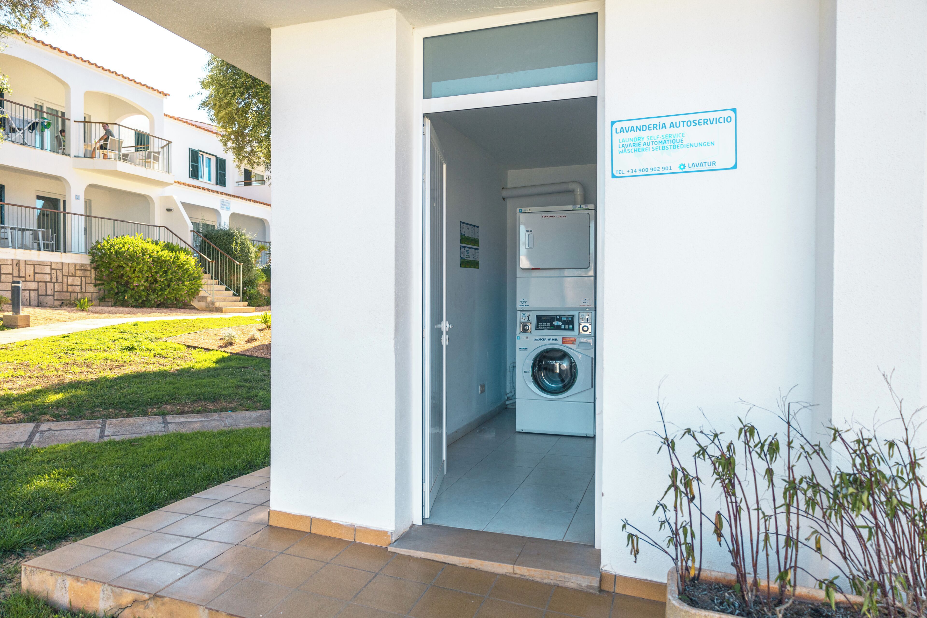 Una lavanderia con lavatrice e asciugatrice, un cartello che indica servizio di lavaggio self-service, e sullo sfondo un edificio residenziale.