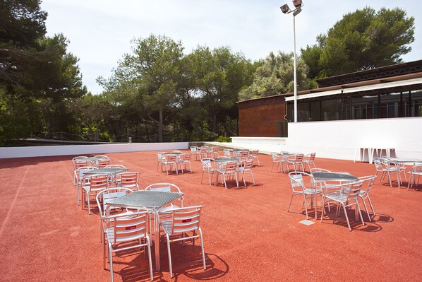 Terraza al aire libre con varias mesas y sillas, rodeada de árboles y un edificio con grandes ventanales.