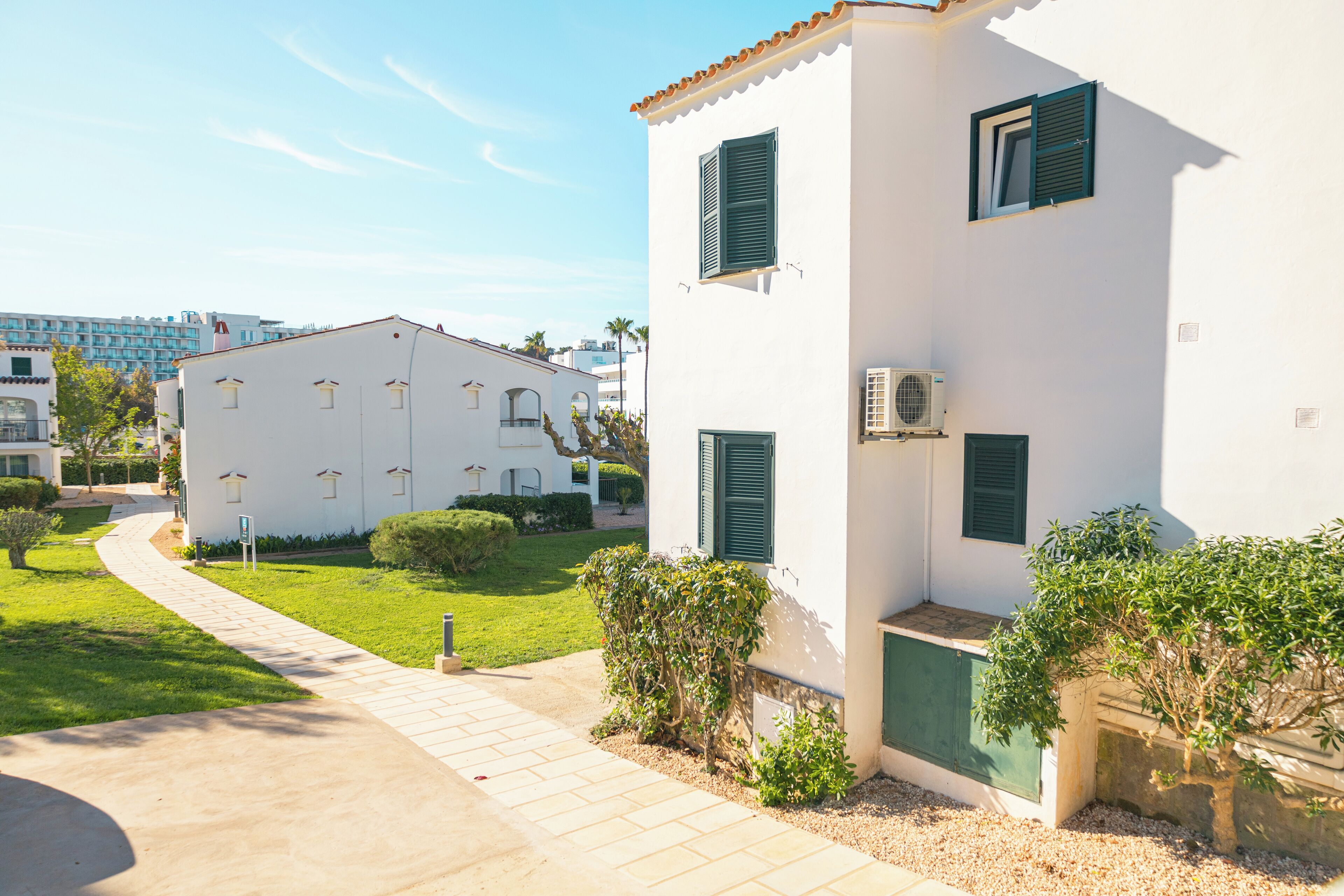 Un edificio blanco con contraventanas verdes, un camino de entrada pavimentado y un pequeño jardín.