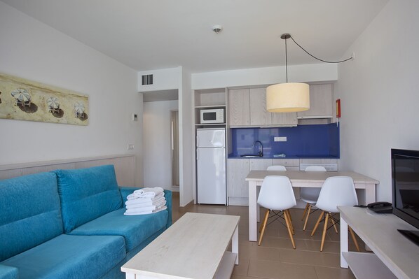 A modern living room with a blue sofa, a white coffee table, and a kitchen area with white cabinets, a microwave, and a sink.