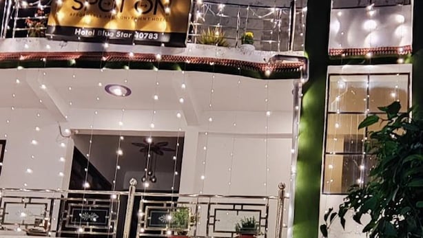 Ein Hotel namens 'Blue Star' mit einem Schild, das Zimmer, ein Restaurant und einen Ballsaal anzeigt. Vor dem Gebäude befinden sich Blumentöpfe und ein Roller.