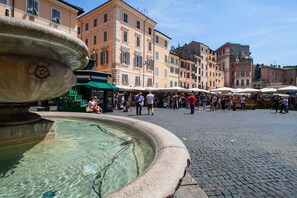 A public square with a fountain, cobblestone pavement, and historic buildings in the background.