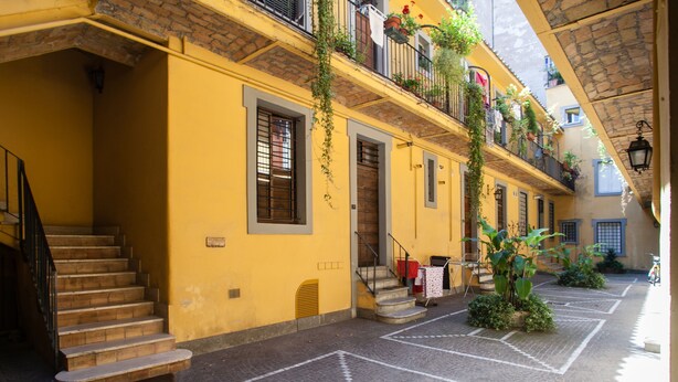 A yellow building with a staircase leading to a second floor. There are plants on the balconies and a tiled floor. - Cozy Apartment in via dei Cappellari, Campo de' Fiori (Rome)