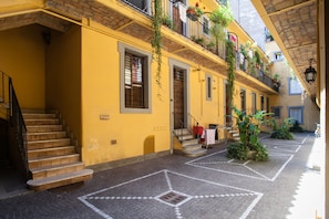 A yellow building with a staircase leading to a second floor. There are plants on the balconies and a tiled floor.