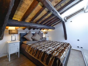 A bedroom with a sloped ceiling, wooden beams, a bed with a fur throw, bedside lamps, and a nightstand. - Ibernesi 1 Apartment (Rome)