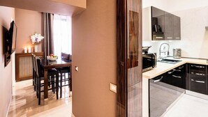 A modern kitchen with dark cabinetry, a countertop, and a sink. There is a dining area with a table and chairs, and a vase with flowers on the wall. - The Brunetti - Luxury Serviced Apartment (Rome)