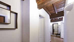 Un pasillo con techo de madera, paredes blancas y piso de baldosas. Hay una puerta al final.