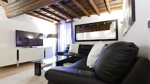 Una sala de estar con un sofá de cuero negro, una mesa de centro de vidrio, un televisor de pantalla plana y un aparador blanco.
