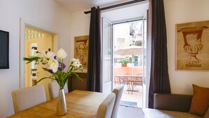 Un comedor con una mesa de madera, sillas blancas y un florero. Hay un ventanal corredero que da a una zona de estar exterior con mesa y sillas.