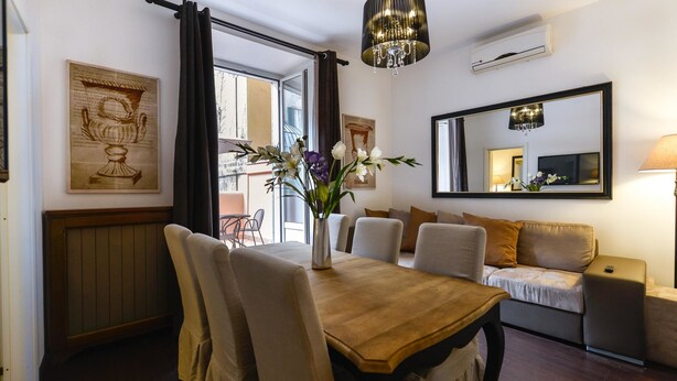 Un comedor con una mesa de madera, sillas, un jarrón con flores, una araña, un sofá, un televisor y un espejo.