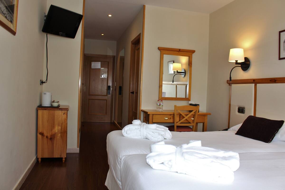 Une chambre d’hôtel avec deux lits, un bureau en bois, un miroir, un téléviseur fixé au mur et une petite armoire.