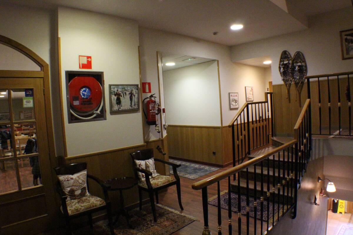 Un hall d’entrée avec un escalier en bois, deux chaises, un extincteur et une horloge au mur.