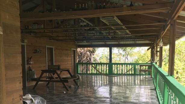A covered outdoor area with a tiled floor, a green railing, and a wooden structure with a metal roof. - Island Charm Apartments (Roatan)