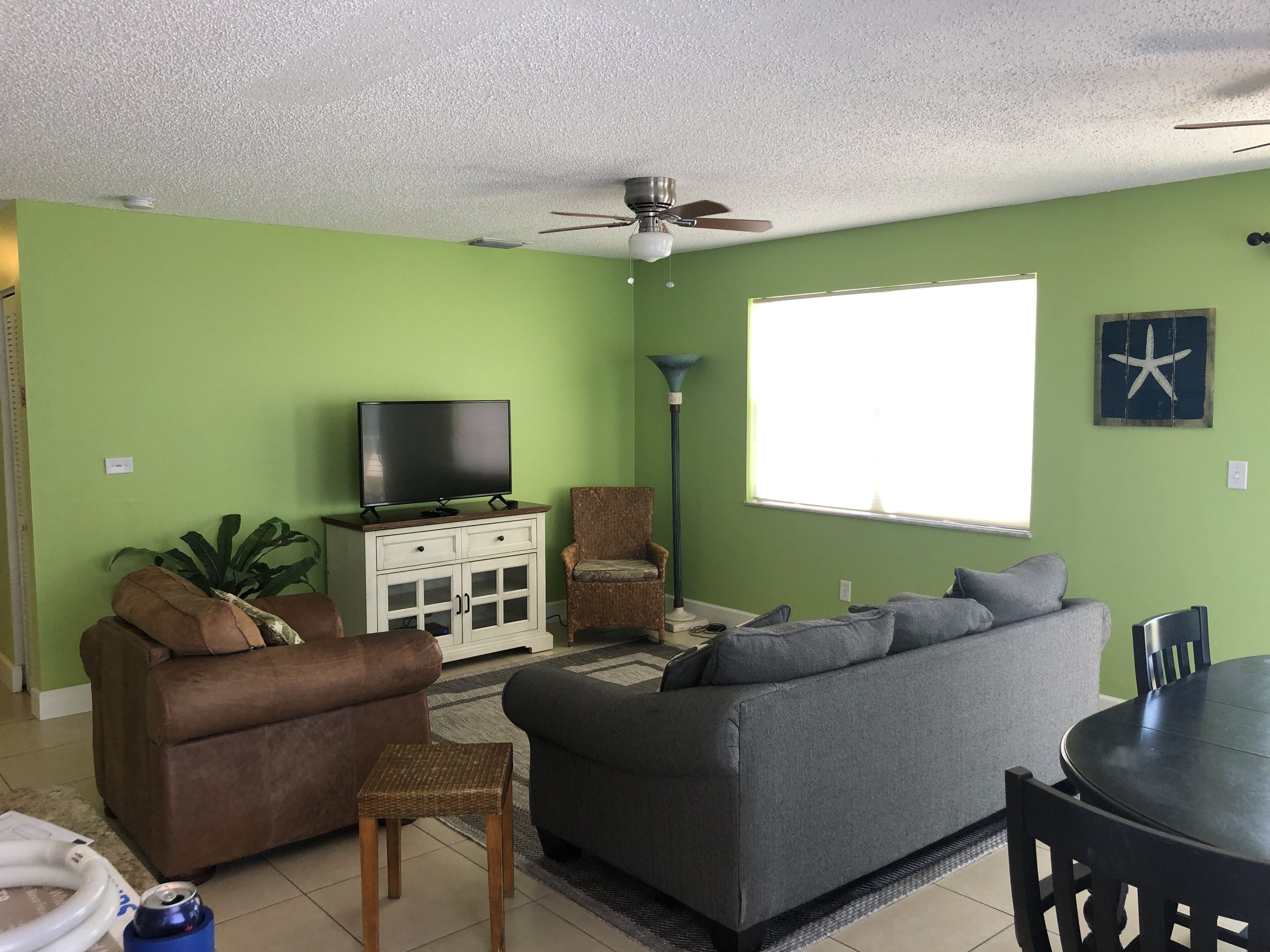 A living room with a green wall, a ceiling fan, a flat-screen TV on a cabinet, a brown armchair, a gray sofa, a round table, and a starfish decoration.