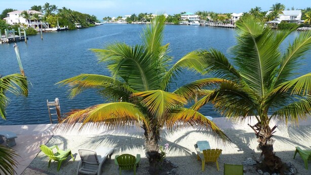 A beachside view with palm trees, a sandy area with chairs, and a body of water with buildings in the background. - 2 Incredible Waterfront Homes - One Low Price! (Marathon)