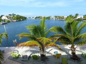 A beachside view with palm trees, a sandy area with chairs, and a body of water with buildings in the background.