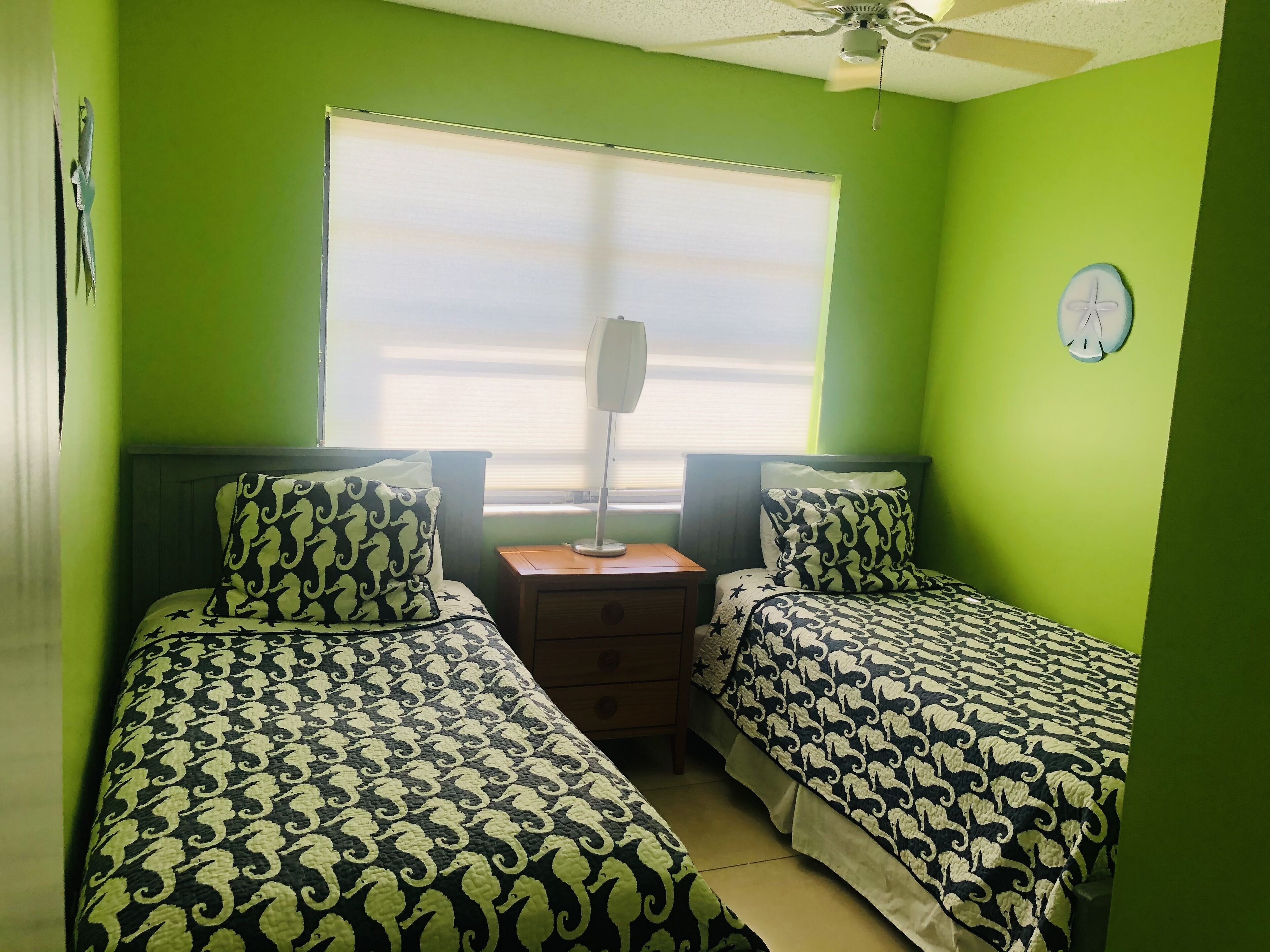 Une chambre avec deux lits, une table de chevet, une lampe, une fenêtre avec des stores et un ventilateur de plafond.