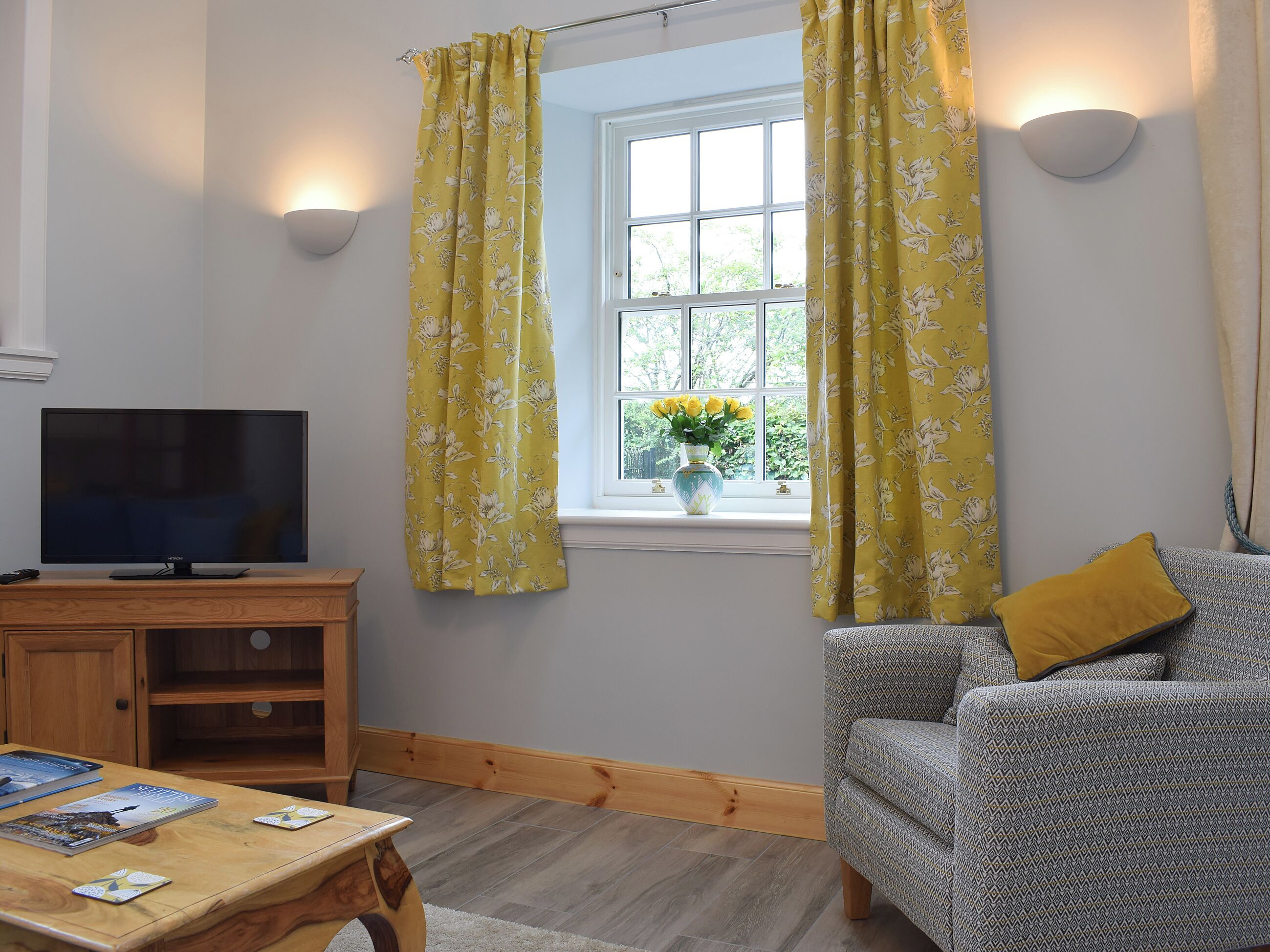 Een woonkamer met een televisie, een houten salontafel, een sofa met een patroon en een raam met bloemen gordijnen in geel.