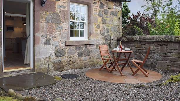 A stone building with a kitchen visible through an open door, a small outdoor seating area with a table and chairs, and a stone wall in the background. - The Old Stables At Westerton (Crieff)