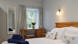 A neatly made bed with white linens, a wooden nightstand with a vase of yellow flowers, a mirror, and a window with checkered curtains. - The Old Stables At Westerton (Crieff)