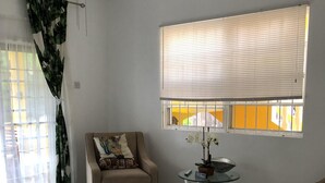 Un ampio soggiorno con ventaglio a soffitto, una poltrona beige, un tavolino in vetro con una pianta e una finestra con persiane.