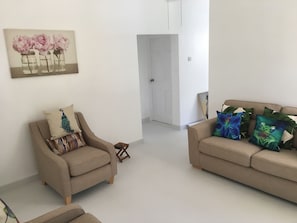 En stue med en beige sofa, en lænestol med pude med påfuglemotiv, og en farverig blomsterpude på sofaen.
