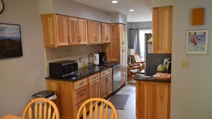 Una cocina con armarios de madera, microondas, hervidor y comedor con sillas.