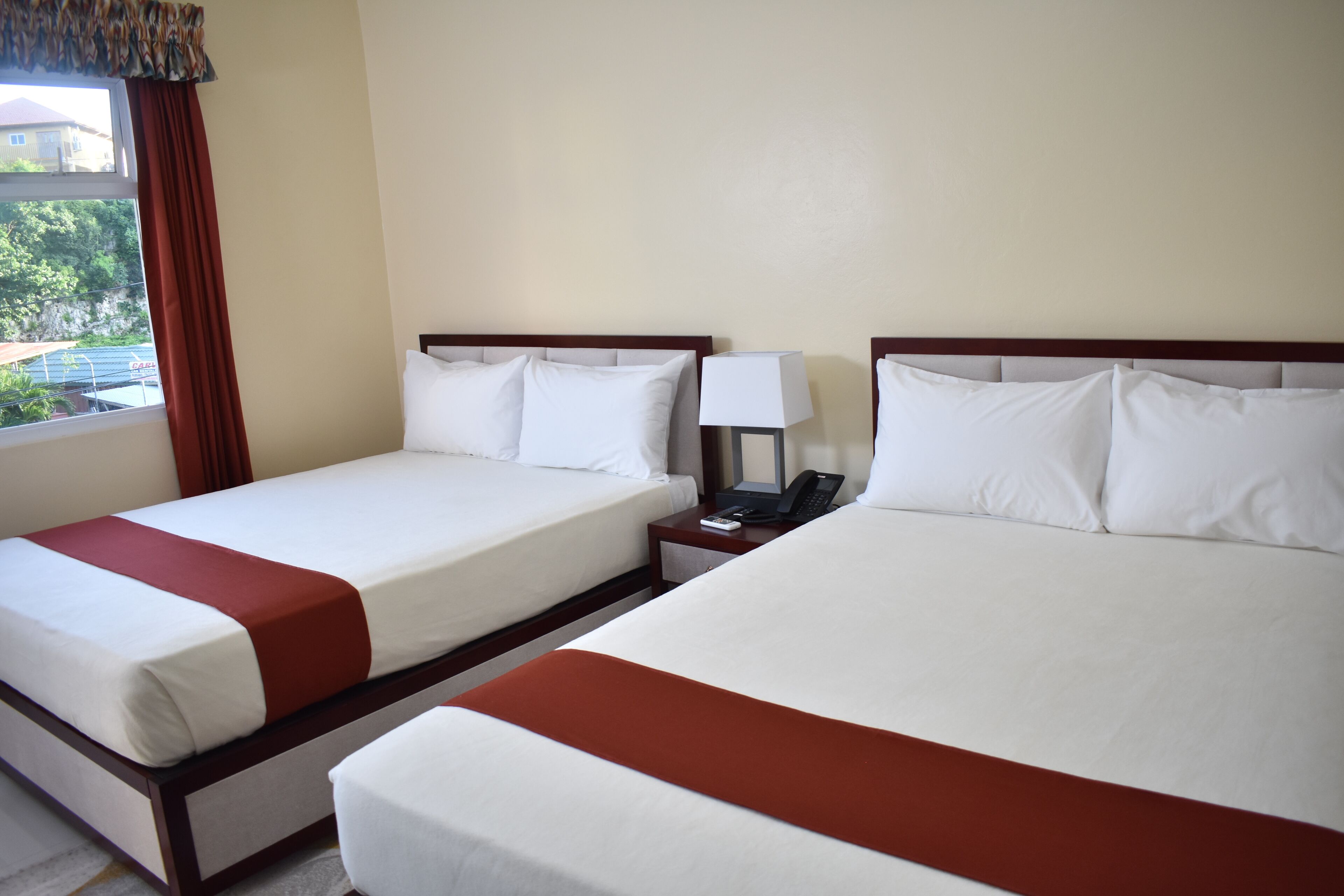 Two beds with white linens and brown accents, a bedside table with a lamp, and a window with a view of greenery.