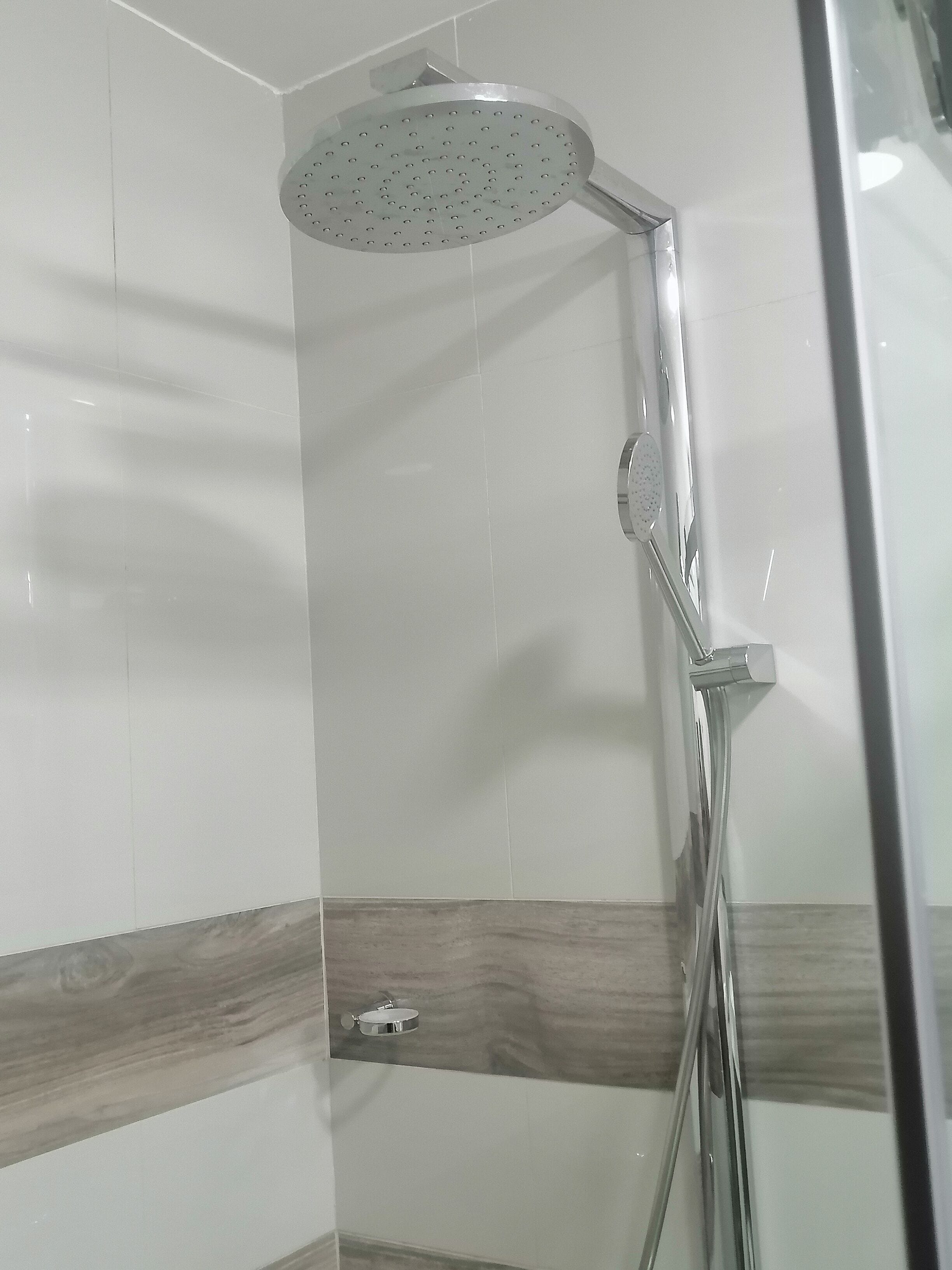 Ducha con cabezal de ducha de lluvia y un cabezal de ducha manual, un perchero y un panel de madera en la pared.