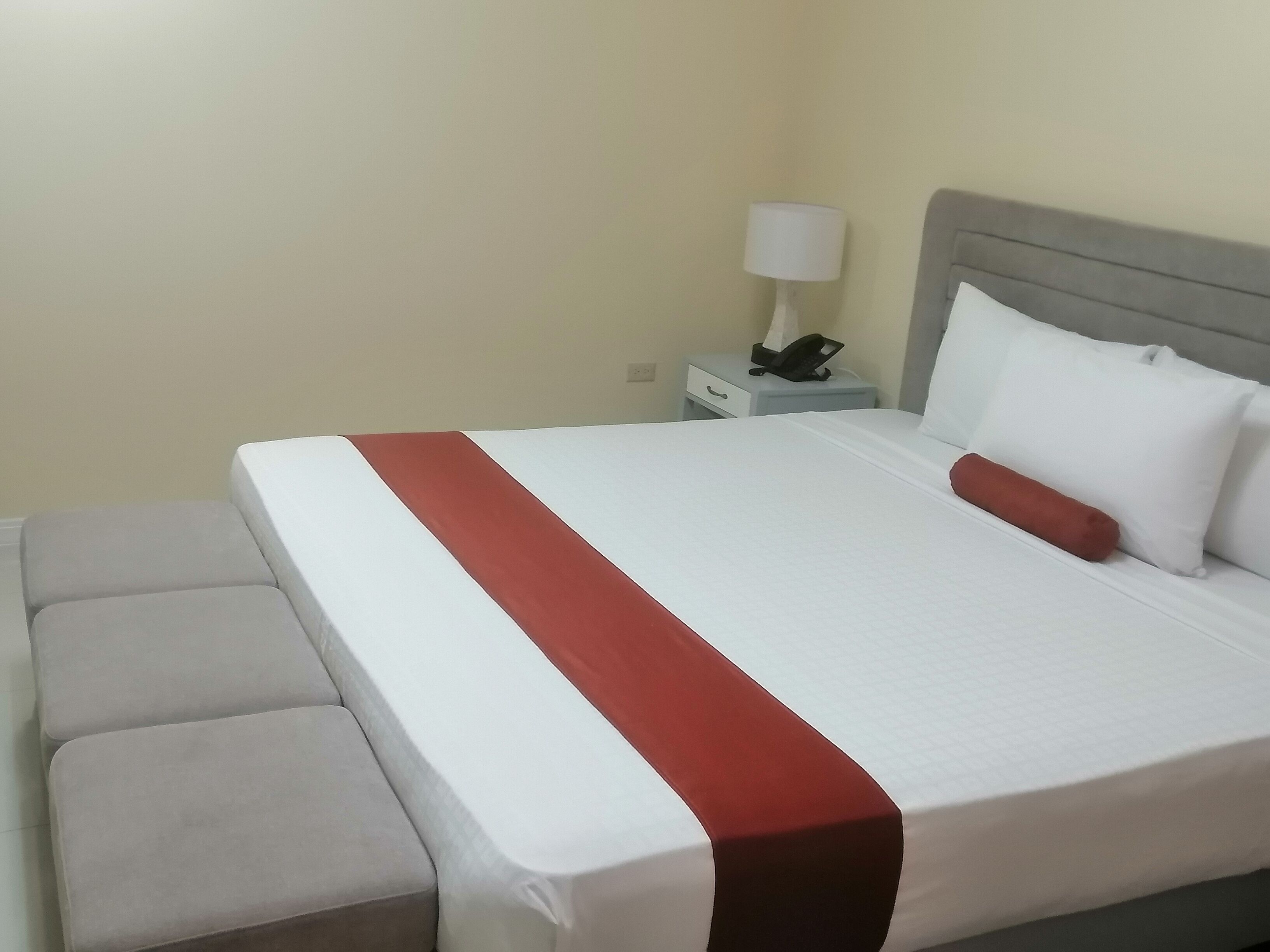 A neatly made bed with a white and red bedspread, a grey headboard, and a bedside table with a lamp and phone.