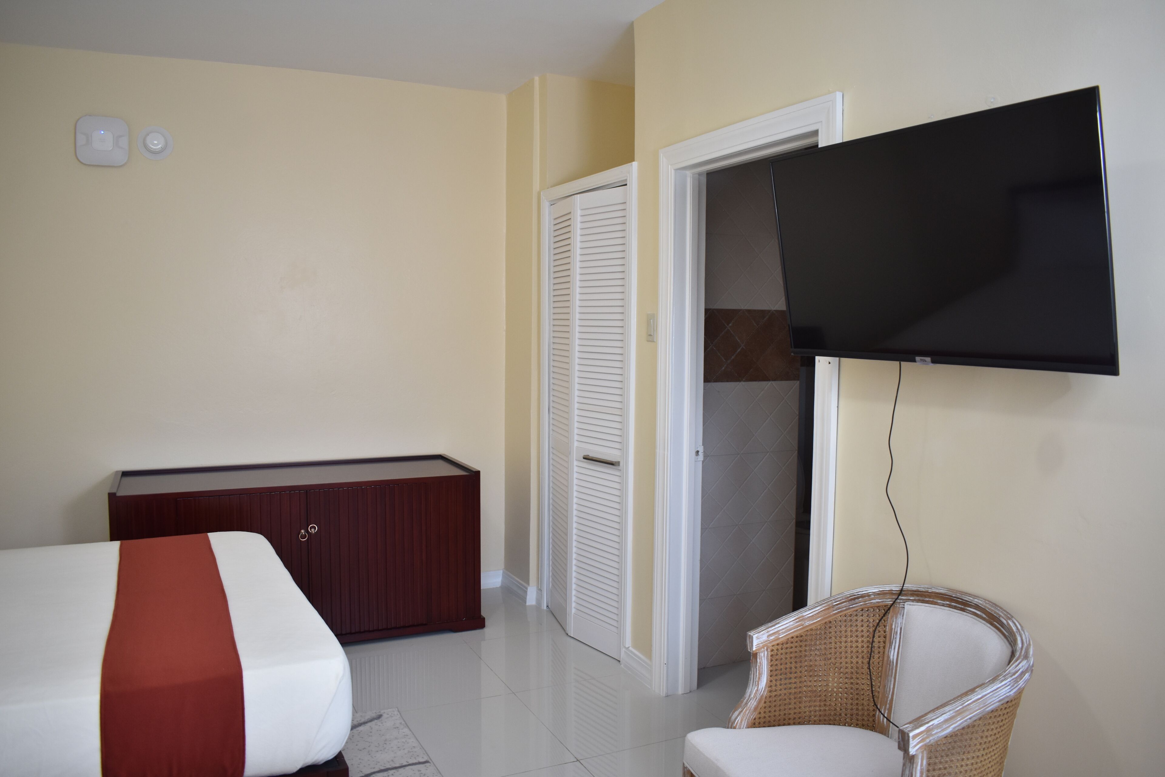 Ein Hotelzimmer mit einem Bett, einer Holzkommode, einem an der Wand befestigten Fernseher und einem Korbstuhl.