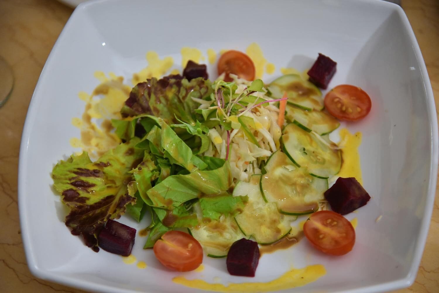 A white square plate with a salad consisting of lettuce, cucumber, tomato, and beetroot slices.