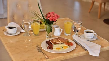 Uma mesa de café da manhã com um prato de ovos, bacon e panquecas, acompanhado de café e suco de laranja.
