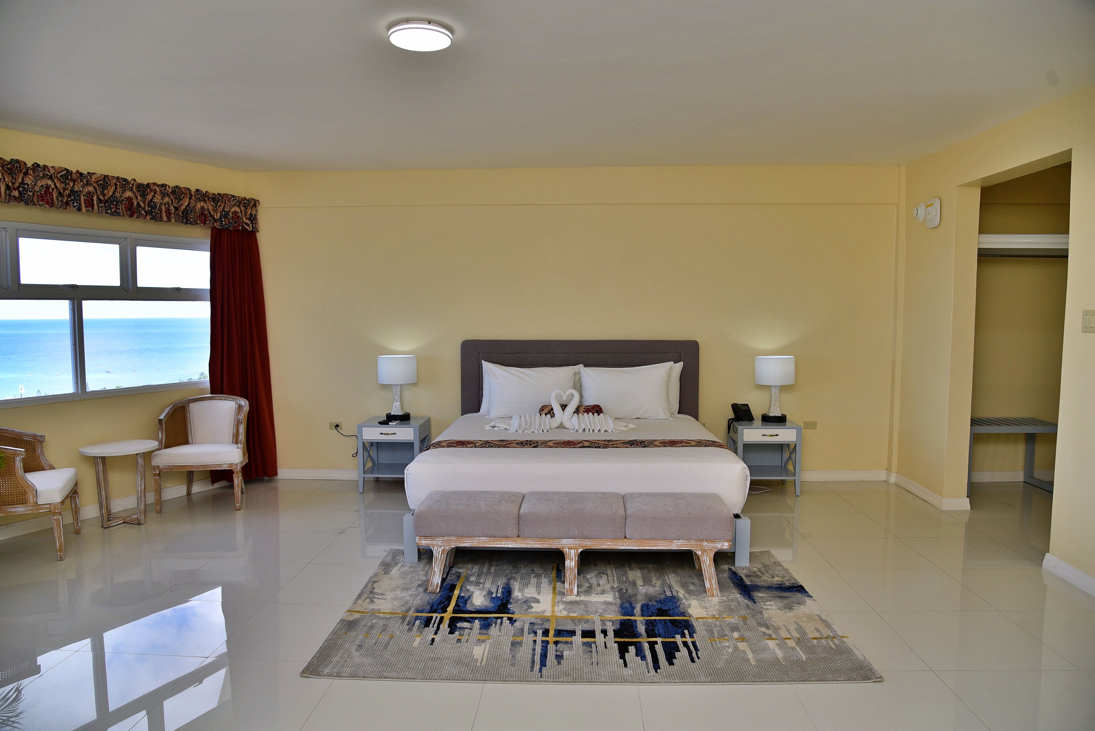 Une chambre d’hôtel avec un grand lit, deux tables de chevet, un banc, une chaise et une vue sur l’océan.