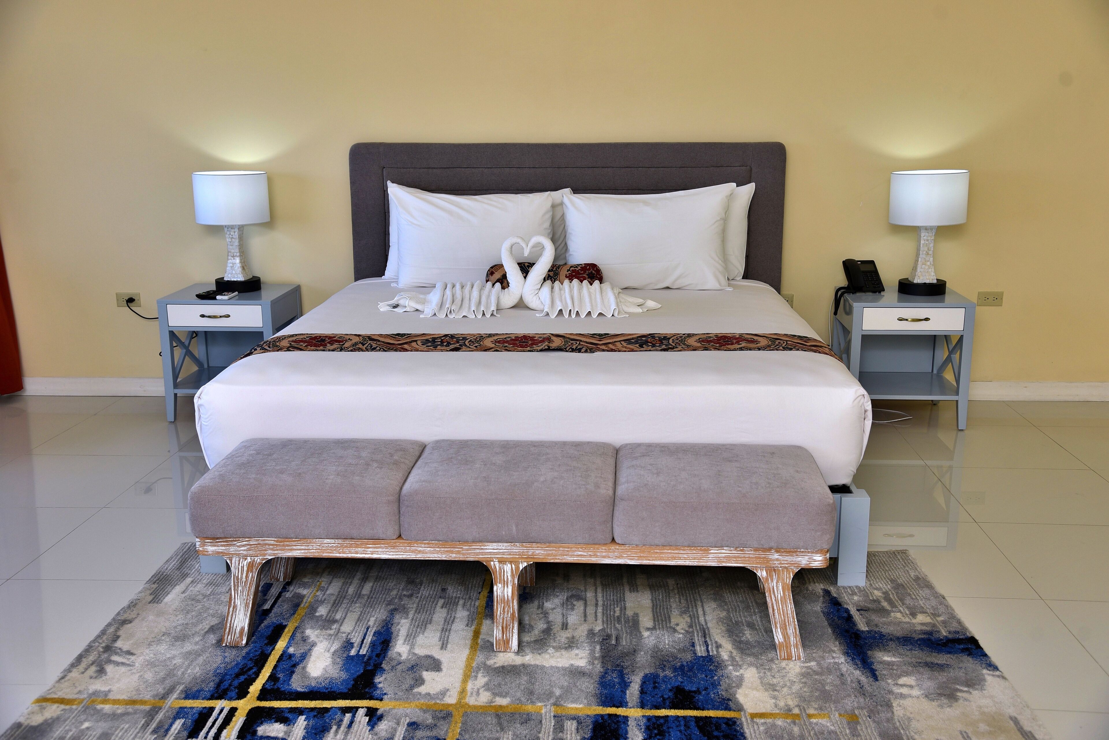 Una cama bien hecha con edredón blanco y almohadas, una mesita de noche con lámpara, y un arreglo decorativo con cisnes sobre la cama.