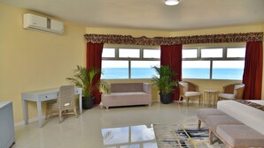 A living room with a sofa, a chair, a small table, and a desk. There are two potted plants and a view of the ocean through large windows. - Hotel Grand A View (Montego Bay)
