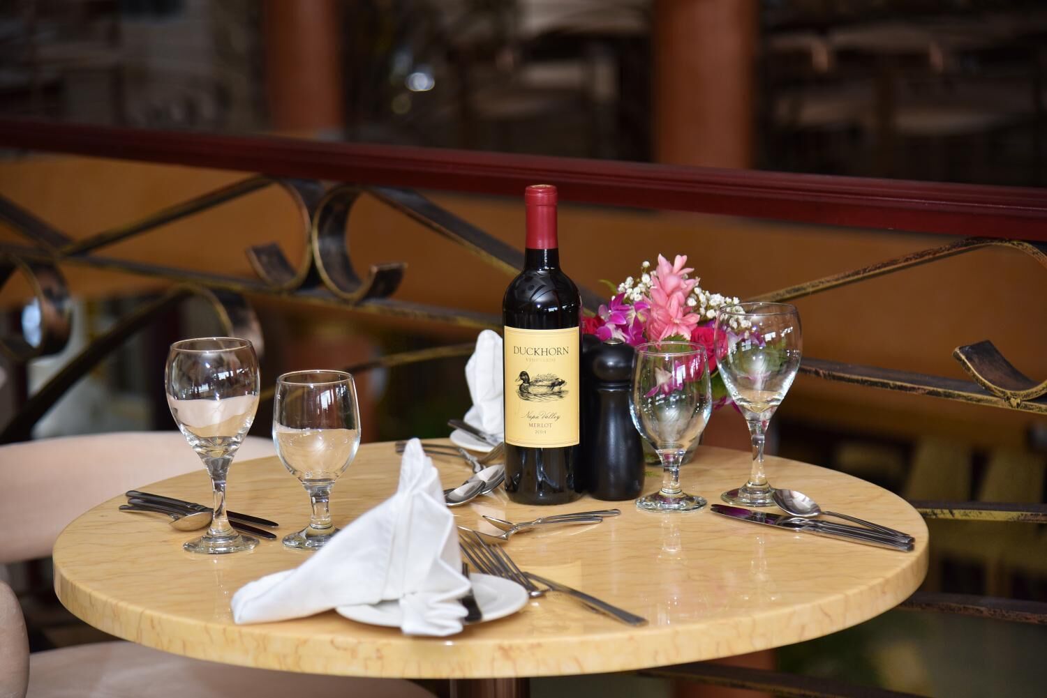 A round wooden table with a bottle of Duckhorn wine, two glasses, a vase with flowers, and a salt and pepper shaker.