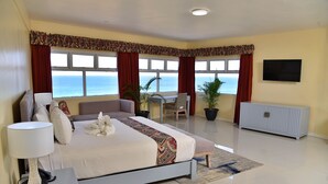 A bedroom with a bed, bedside table, lamp, desk, chair, potted plants, and a view of the sea. - Hotel Grand A View (Montego Bay)