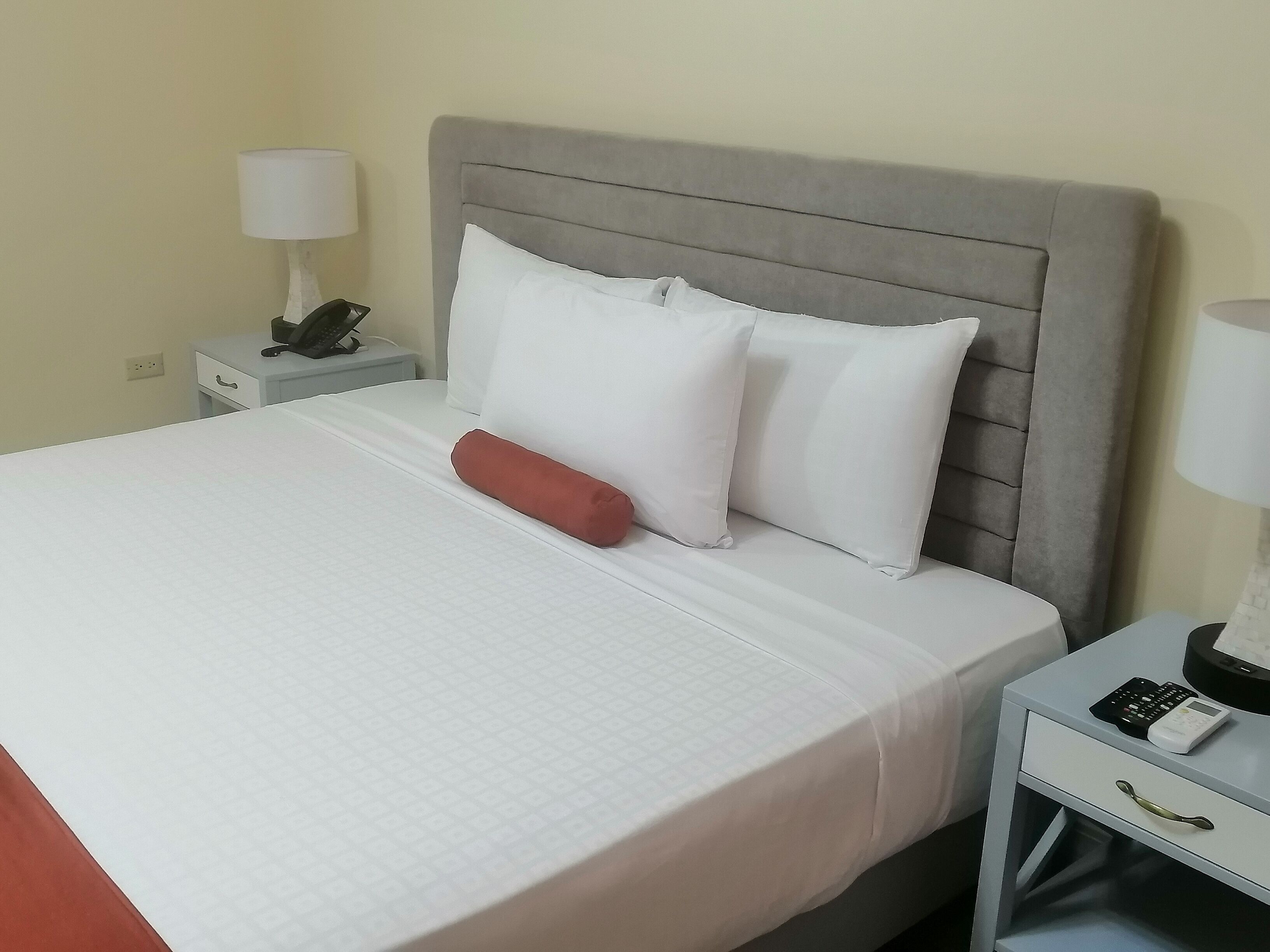 A neatly made bed with white bedding and a grey headboard. There is a red pillow and two white pillows on the bed. A white nightstand with a lamp and a phone is beside the bed.