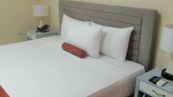 A neatly made bed with white bedding and a grey headboard. There is a red pillow and two white pillows on the bed. A white nightstand with a lamp and a phone is beside the bed.