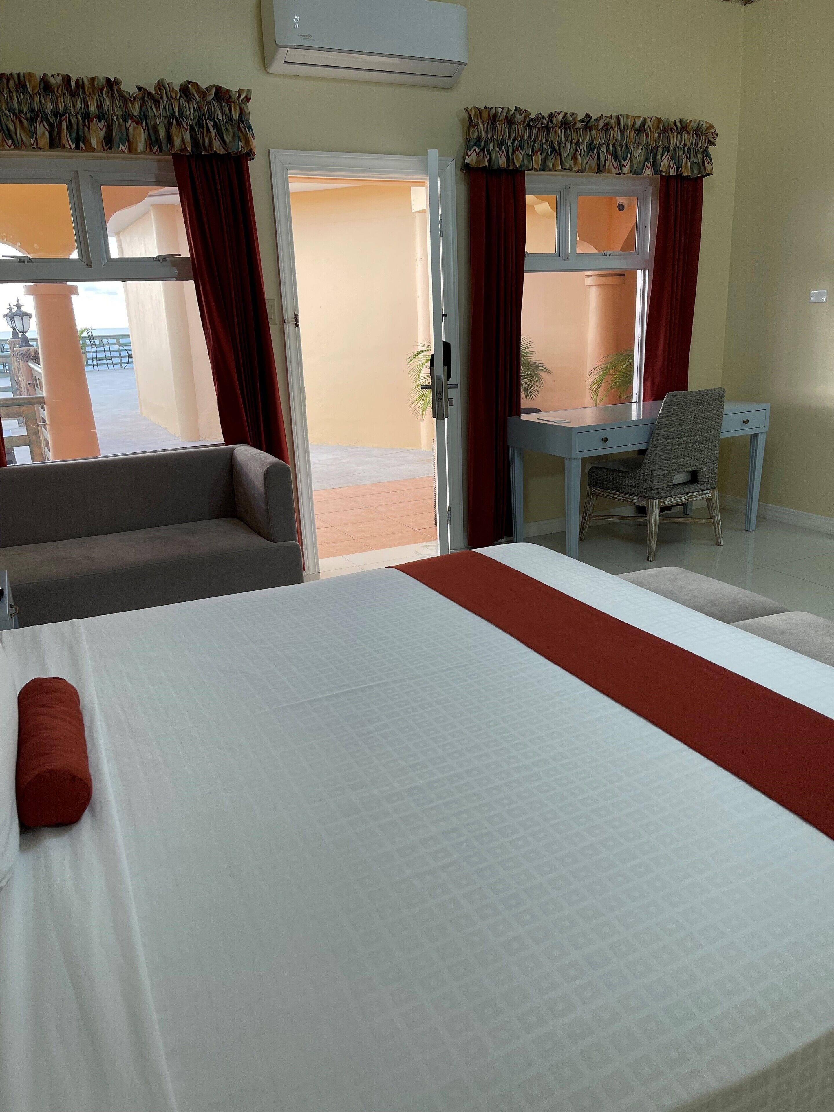 A hotel room with a bed, a desk, and a chair. There is a window with curtains, and an air conditioning unit above it.
