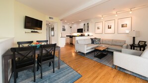 A modern living room with a dining area, a grey sofa, a flat-screen TV, and a kitchen in the background. - Beachfront, East End, 2 BR, 2BA, Private Deck, Parking, Dog Friendly (Provincetown)