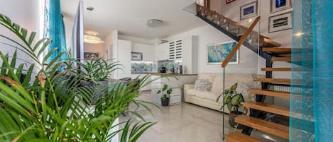Un salon moderne avec un escalier, une cuisine en arrière-plan et diverses plantes.