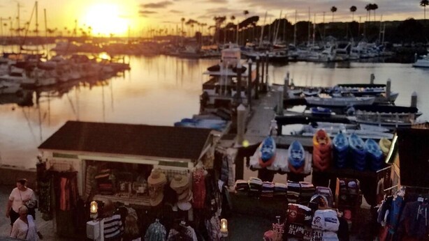 A marina with boats docked, a small shop with various items for sale, and a sunset in the background.