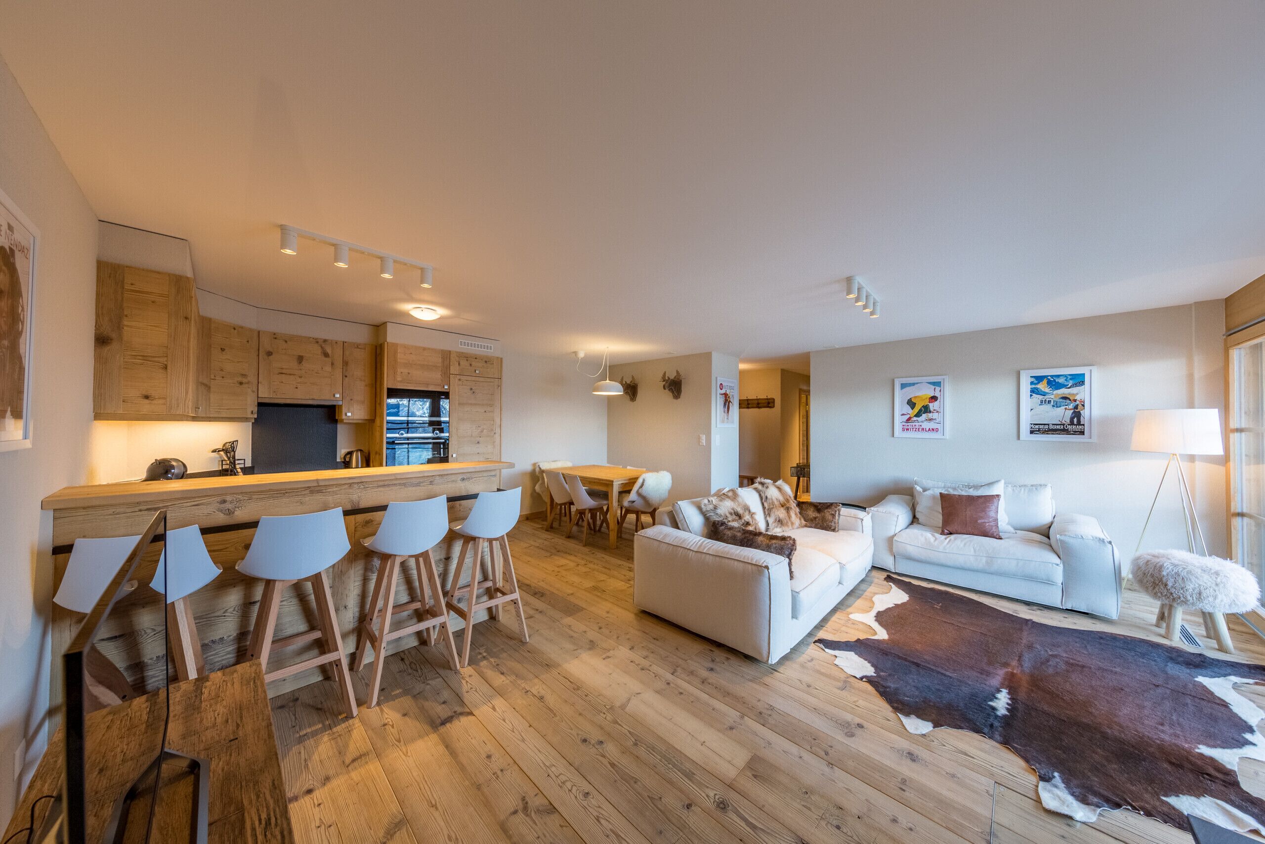 Een moderne keuken met houten kastjes, een bar voorzien van witte barkrukken, een eettafel en een zithoek met een witte bank en een groot koperen vloerkleed.