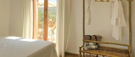 A bedroom with a bed, a window with curtains, a wooden rack with clothes and baskets, and a view of the outdoors.