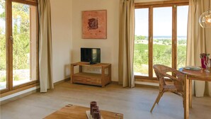 A room with a TV on a wooden stand, a dining table with chairs, and large windows offering a view of greenery. - Lovely home in Son Carrió with WiFi (Son Carrió)